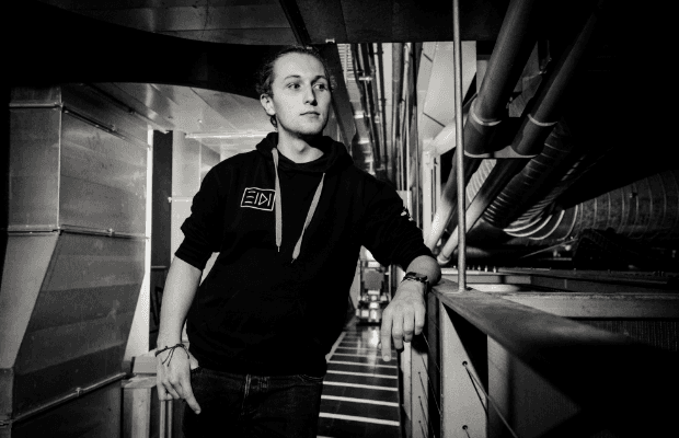 DJ Eidi ist frontal zu sehen. Er trägt einen schwarzen Kapuzenpullover und blickt nach links. Seinen linken Arm stützt er lässig auf ein Geländer. Das Foto ist schwarz-weiß.