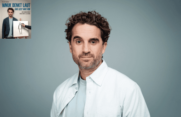 Porträt des Schauspielers und Autors Oliver Wnuk, mit dunklen lockigen Haaren, braunen Augen und Dreitagebart, der vor einem graublauen Hintergrund stehend légère ein weißes, geöffnetes Hemd über einem hellblauen T-Shirt trägt und entspannt in die Kamera blickt.