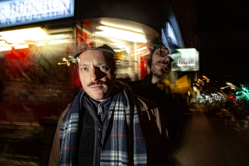 Zwei Musiker stehen nachts auf einer urbanen Straße, bewegt aufgenommen mit Lichtspuren im Hintergrund.