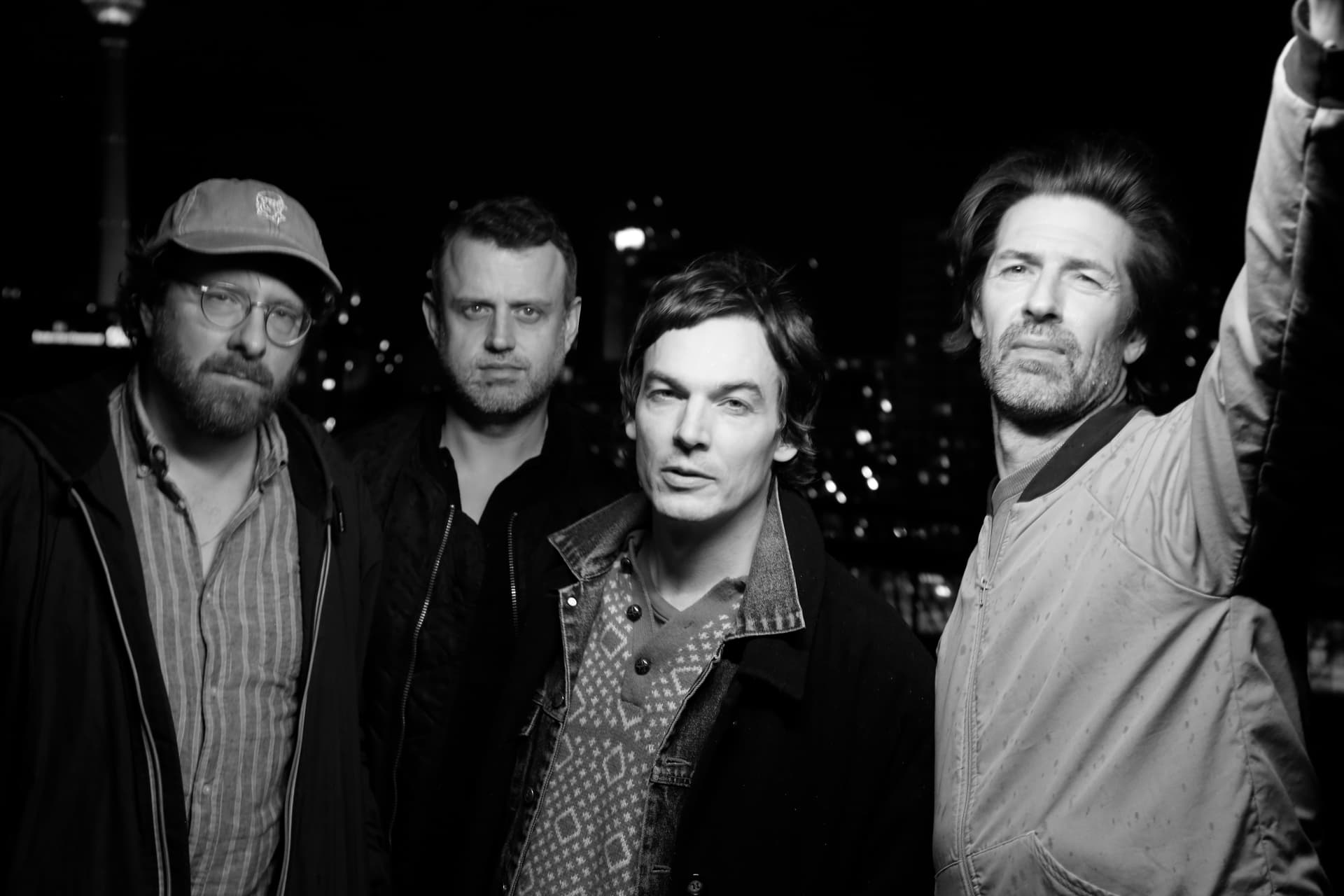 Die vier Musiker blicken ernst in die Kamera. Die Person rechts im Bild streckt den linken Arm in die Höhe. Das Bild ist schwarz-weiß.