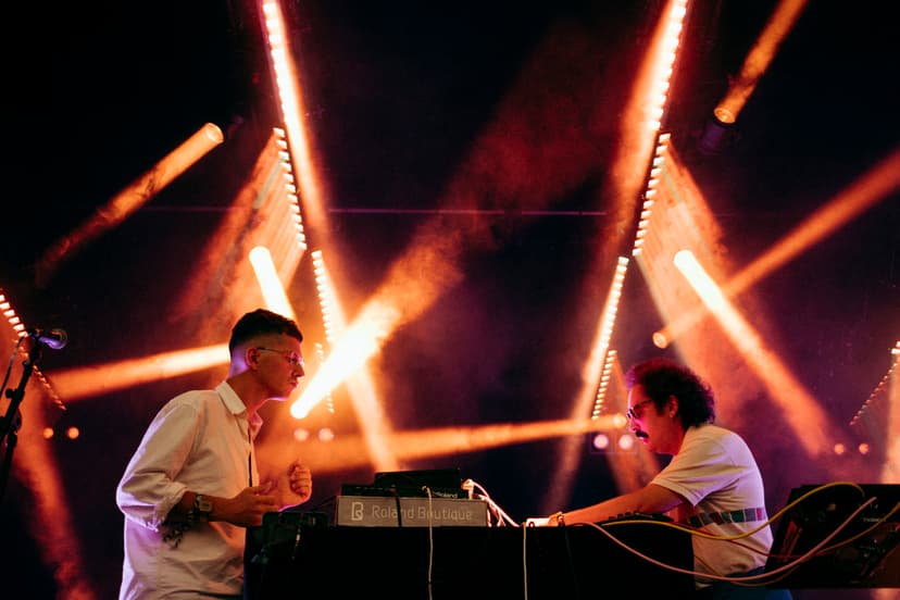 Die Musiker des Bandprojekts Mind Enterprises stehen auf einer Bühne an Synthesizern und elektronischem Equipment, umgeben von orangefarbenen Lichtstrahlen und Nebel.