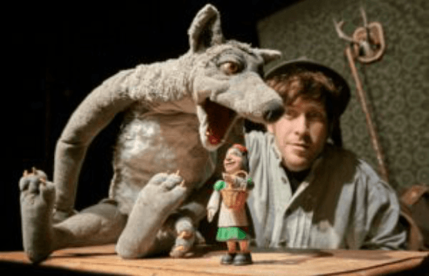 Ein Mann mit Hut und zwei Puppen: Ein Wolf der sein Maul über die kleine Figur eines Mädchens mit roter Kappe beugt.