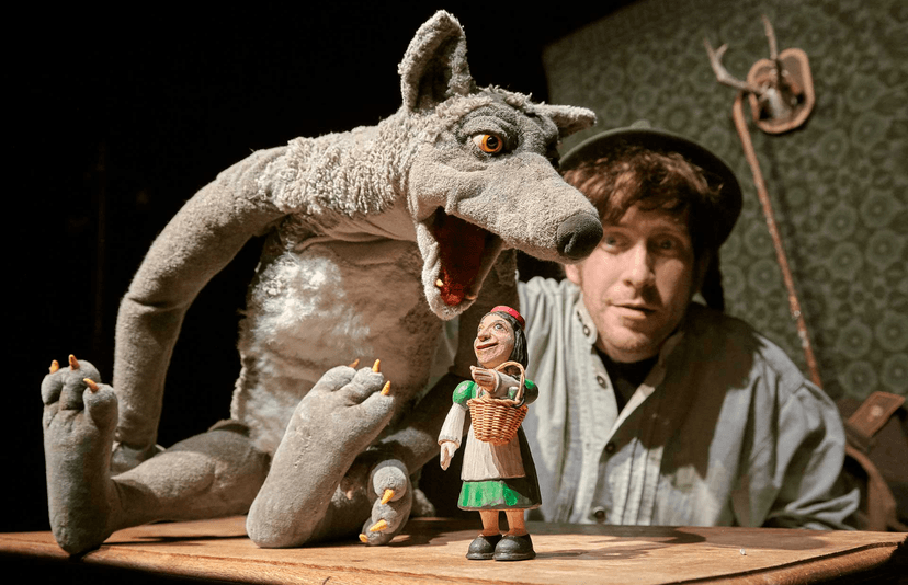 Ein Mann mit Hut und zwei Puppen: Ein Wolf der sein Maul über die kleine Figur eines Mädchens mit roter Kappe beugt.