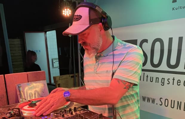 DJ Katz Records steht am Mischpult und scratcht eine Schallplatte. Er trägt eine Schirmmütze und große Kopfhörer.