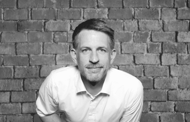 Schwarz/Weiß-Fotografie von DJ Daniel Shy, der vor einer gemauerten Wand steht, ein lockeres weißes Hemd trägt und lächelnd in die Kamera blickt.