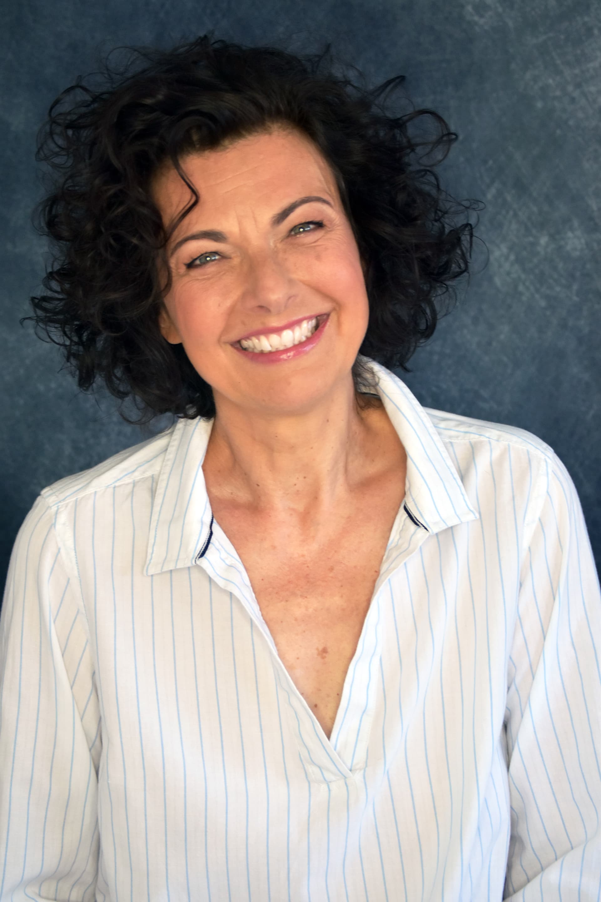 Porträt der Autorin Antje Herden mit dunklen, lockigen Haaren vor blauem Hintergrund, die lächelnden in die Kamera blickt.