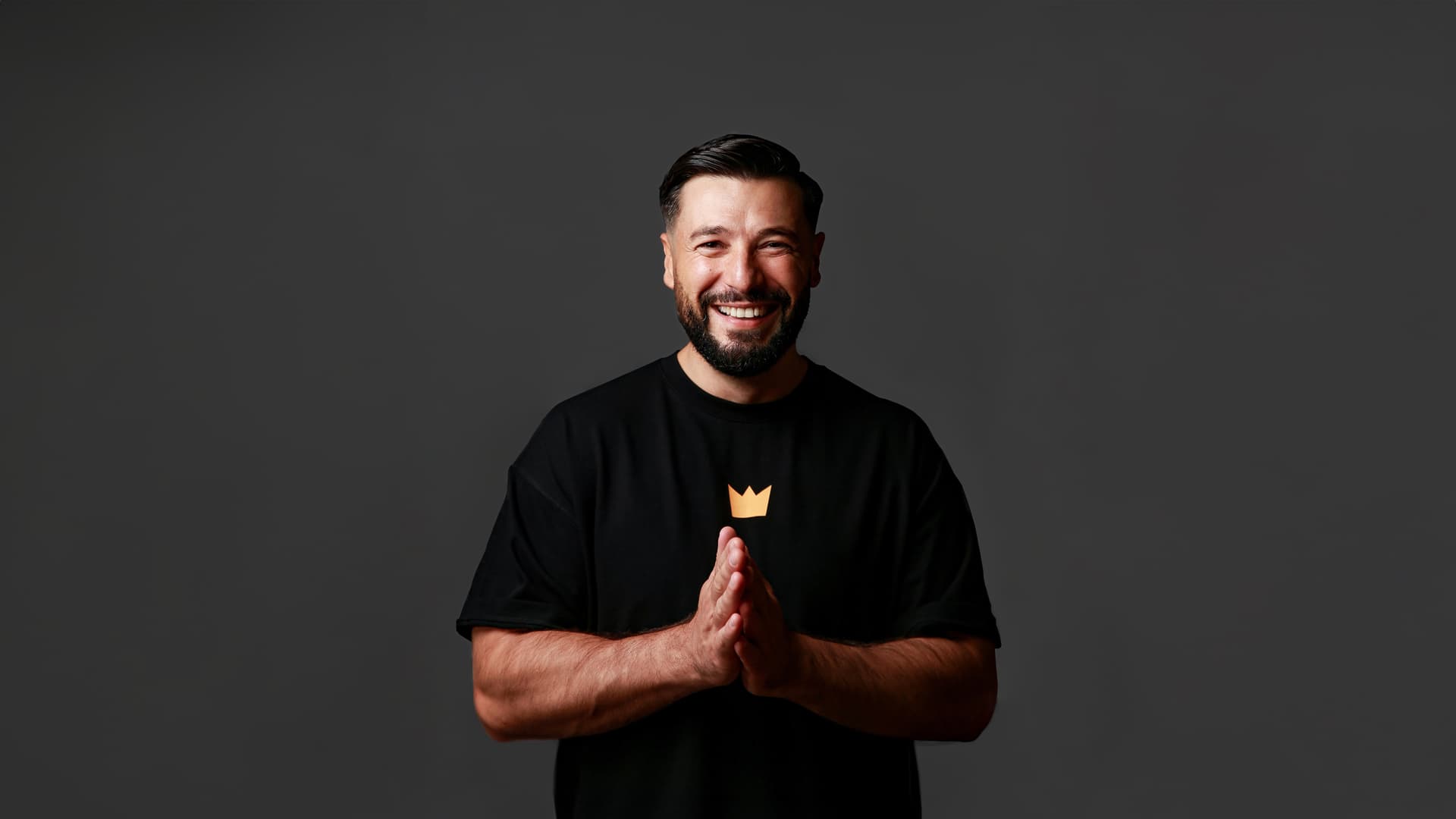 Comedian Cüneyt Akan lächelt vor dunklem Hintergrund in die Kamera, hält die Hände vor der Brust zusammen und trägt ein schwarzes T-Shirt mit Kronensymbol.