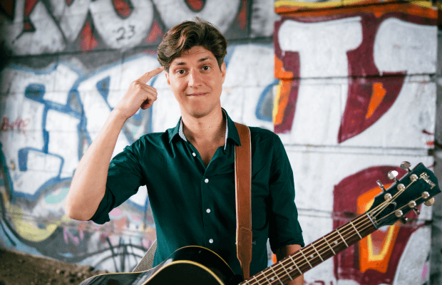 Der Musiker Falk trägt eine Gitarre am Schultergurt und zeigt sich mit dem rechten Zeigefinger an die Stirn. Er steht vor einer Graffiti-Wand.