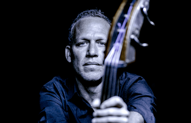 Porträt des Jazzmusikers Avishai Cohen, der frontal in die Kamera blickt und einen Kontrabass hält; das Instrument verläuft unscharf im Vordergrund, sein Gesicht ist kühl ausgeleuchtet vor dunklem Hintergrund und vermittelt eine konzentrierte, ernste Stimmung.