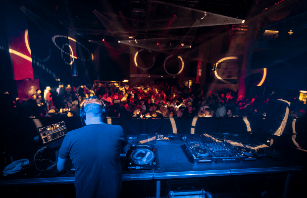 Der DJ steht mit dem Rücken zum Betrachter im Bild. Im Hintergrund sind tanzende Menschen zu sehen, die von buntem Licht angestrahlt werden.