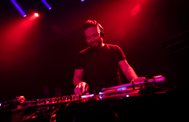 Ein DJ steht am Mischpult und dreht an den Reglern, das Licht ist rot, zwei blaue Strahlen scheinen im Hintergrund.