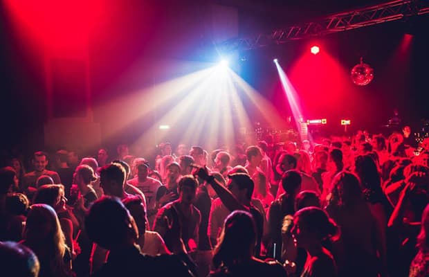 Es ist eine Partyszene in rotem Licht zu sehen. Viele Menschen sind auf der Tanzfläche und werden von einem hellen Strahler beleuchtet. Im Hintergrund hängt eine Discokugel an der Decke.