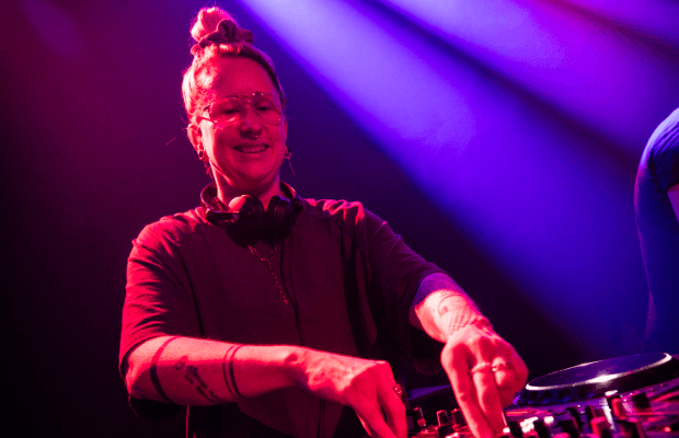 Zu sehen ist eine junge Frau, die mit beiden Händen ein DJ-Mischpult bedient. Das Licht ist in Rot- und Blautönen gehalten, im Hintergrund sind Lichtstrahlen erkennbar.