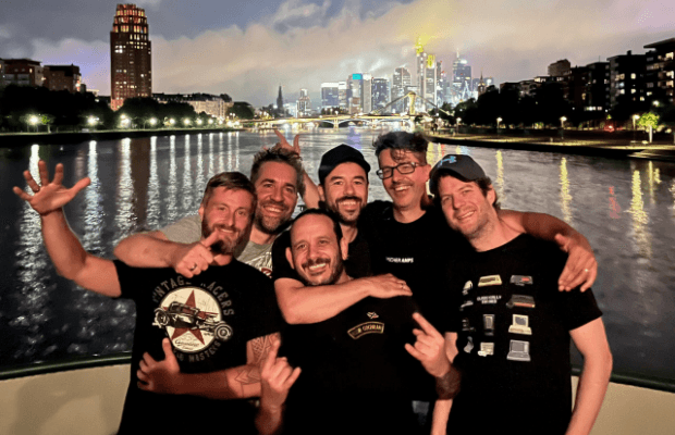 Fröhliches Gruppenfoto von sechs Männern in schwarzen T-Shirts die am Bug eines Schiffs stehen und in die Kamera posieren. Im Hintergrund sind der Fluss sowie die erleuchtete Skyline von Frankfurt am Main bei Nacht zu sehen.