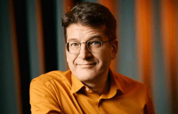 Porträt des Comedians Nils Heinrich in orangenem Hemd und mit Brille, der freundlich in die Kamera blickt.