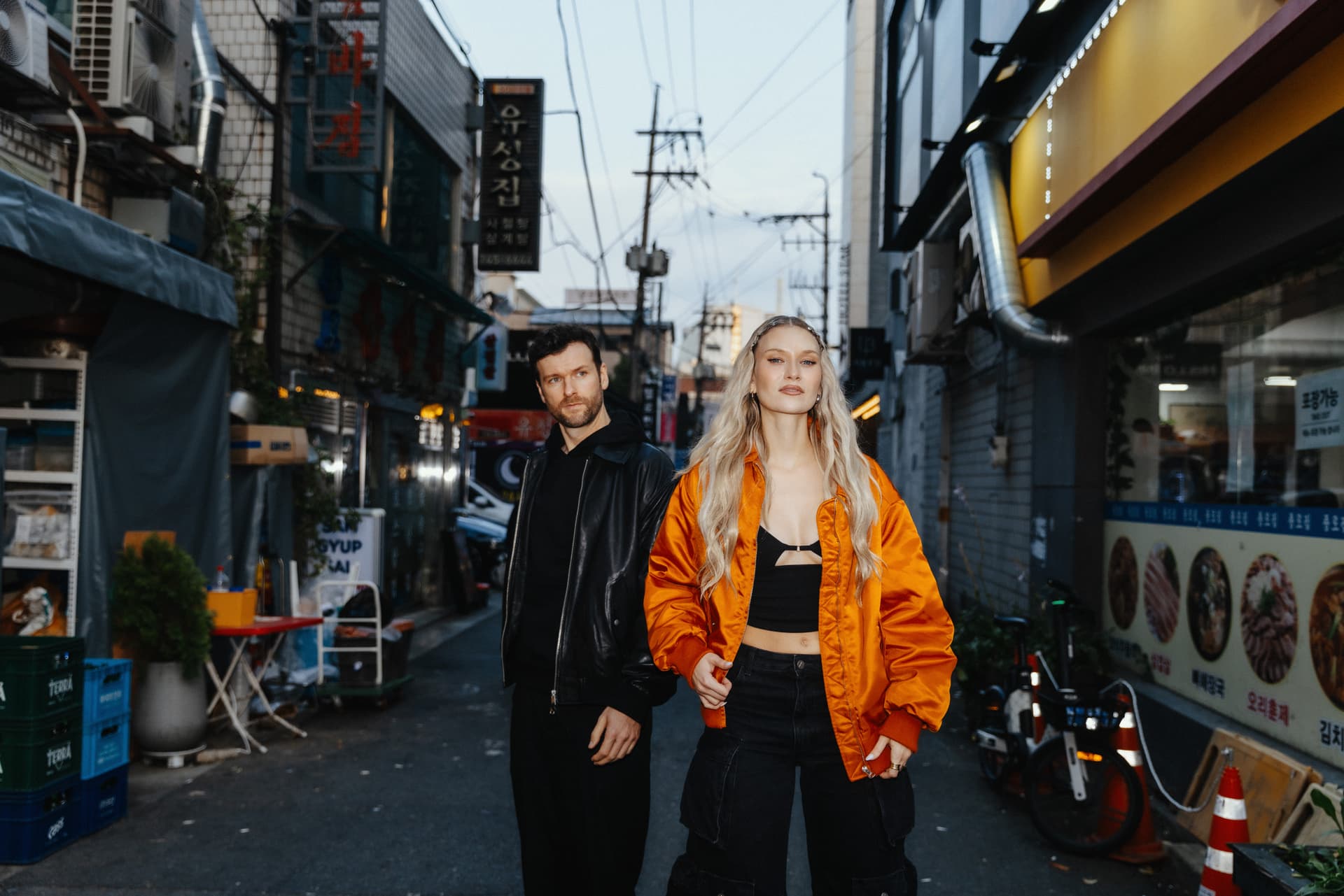 Die Musiker der Band stehen in einer schmalen Straße mit Geschäften, Schildern und Lichtern. Carolin rechts im Bild trägt eine leuchtend orangene Bomberjacke, Daniel links von ihr ist ganz in schwarz gekleidet.