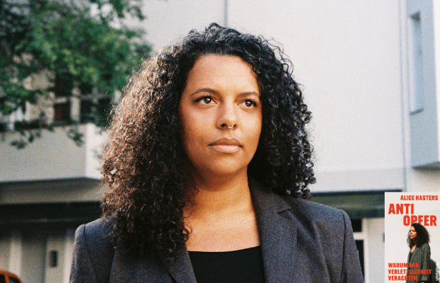 Porträt der Autorin Alice Hasters, im Freien aufgenommen vor einer urbanen Hausfassade. Sie blickt nicht in die Kamera sondern schräg daran vorbei in die Ferne.