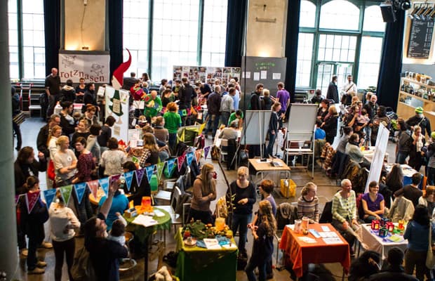 Zu sehen ist eine Ausstellung mit vielen Tischen und Info-Wänden. Es sind viele Personen anwesend und die Halle ist bunt geschmückt.