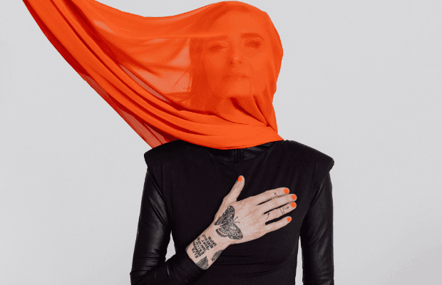 Sarah Lesch in einem schwarzen Outfit mit orangefarbenem Tuch, das über ihren Kopf weht, während sie eine Hand auf ihre Brust legt.