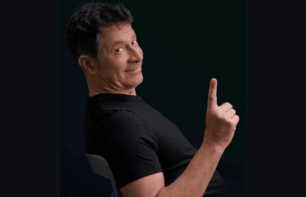 Lächelnder Mann in dunklem T-Shirt seitlich zur Kamera positioniert, zeigt mit erhobenen Zeigefinder nach oben und blickt augenzwinkernd in die Kamera.