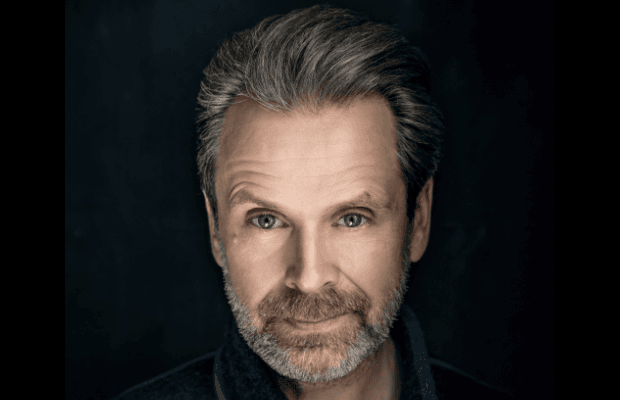 Porträt des Schauspielers Matthias Matschke, der vor dunklem Hintergrund leicht lächelnd in die Kamera blickt.