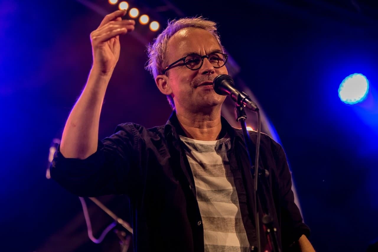 Henni Nachtsheim steht am Mikrofon und macht eine schnipsende Handbewegung in Richtung Kamera. Der Hintergrund ist blau.