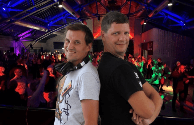 Die DJs der Two Men Show stehen Rücken an Rücken und blicken lächelnd seitlich in die Kamera. Im Hintergrund ist der Saal der Centralstation mit Diskolicht und tanzenden Menschen zu sehen.
