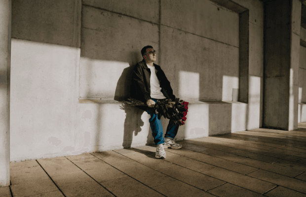 Der Rapper sitzt auf einem Wandvorsprung und hält einen Strauß roter Rosen in der Hand. Die Szene ist in ein Spiel von Licht und Schatten getaucht.