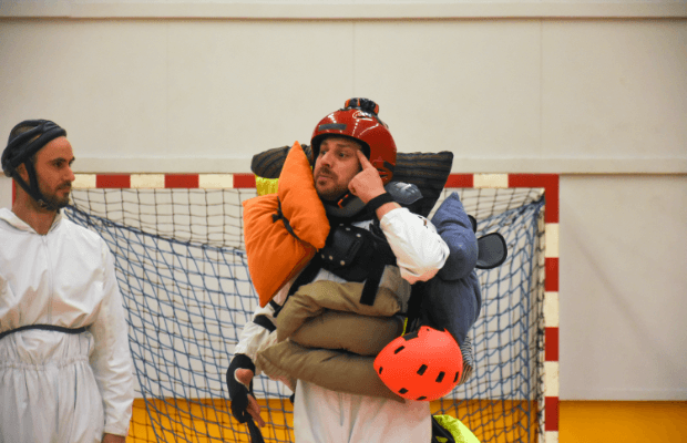Theaterszene in einer Sporthalle: Zwei Männer in Helmen und weißen Overalls, einer davon mit mit Kissen und Polstern behängt.