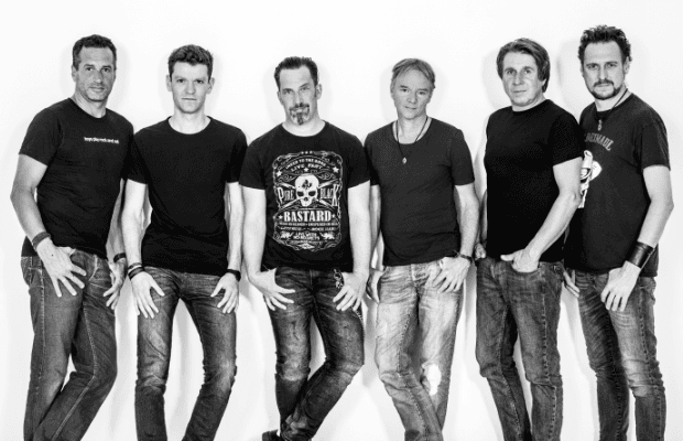Schwarz-weiß-Fotografie von sechs Männer, alle in Jeans und mit schwarzen T-shirts bekleidet, in lässigen Posen vor weißem Hintergrund aufgenommen.