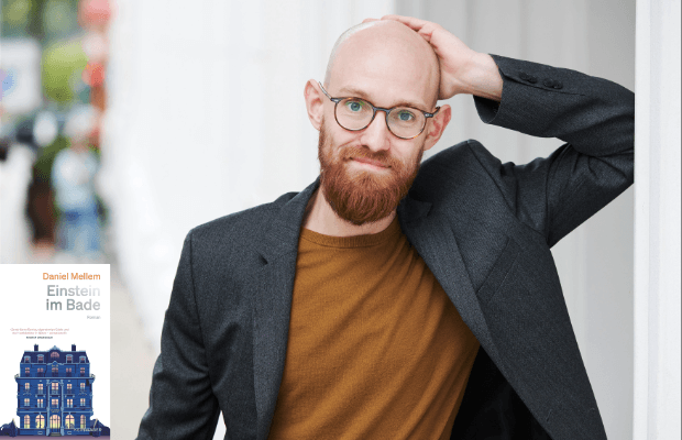 Porträt des Autors mit Vollbart und Brille, er trägt ein braunes Shirt und ein dunkelgraues Sakko darüber und fasst sich mit der linken Hand an den glatt rasierten Kopf.