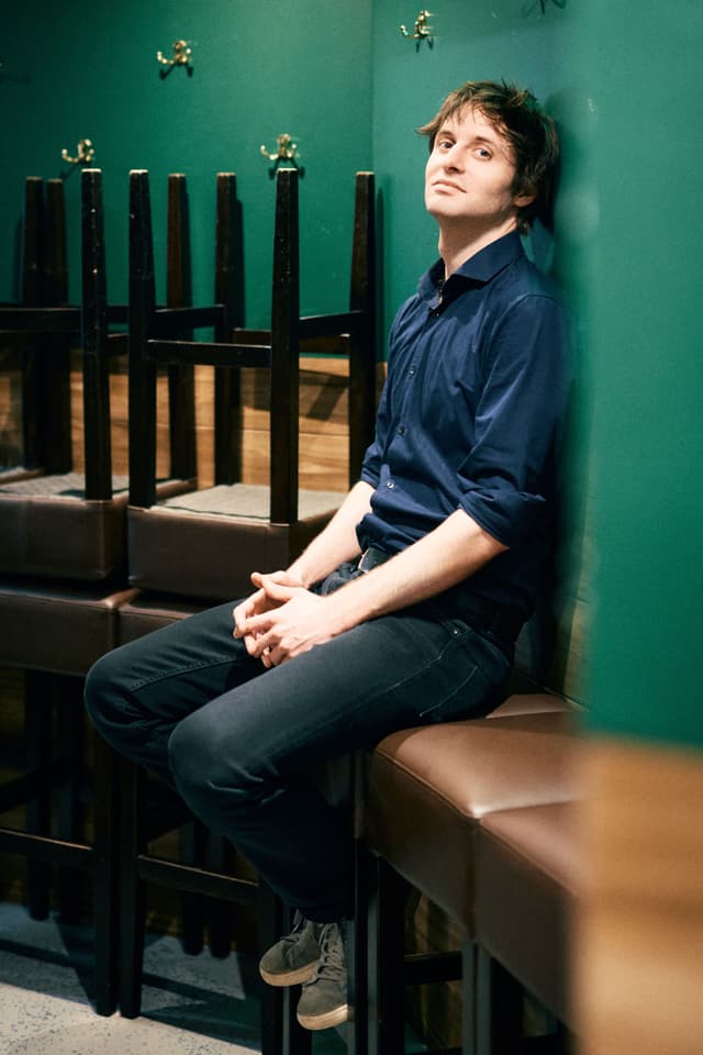 Lennart Schilgen sitzt in einem grün gestrichenen Raum an die Wand gelehnt, neben übereinander gestapelten Stühlen, und blickt ruhig in die Kamera.