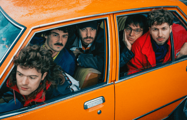 Fünf Musiker der Band Von Wegen Lisbeth sitzen dicht gedrängt in einem orangefarbenen Auto und schauen durch die runtergelassenen Seitenfenster nach draußen.