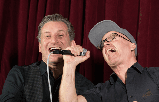 Zwei Männer singen gemeinsam mit einem Mikrofon vor rotem Bühnenvorhang und wirken ausgelassen - Szene vom Rudelsingen.