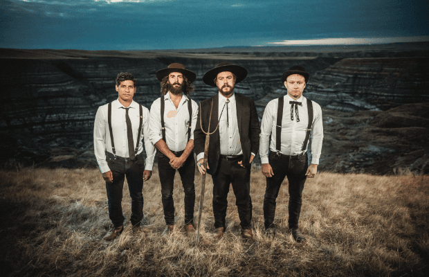 Die vier Musiker stehen nebeneinander in einer weiten, kargen Landschaft, tragen helle Hemden, dunkle Hosen, Hosenträger und Hüte und blicken ernst in die Kamera.