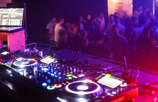 Foto eines Mischpults, farblich gehalten in Blau- und Rottönen. Im Hintergrund sind tanzende Menschen erkennbar.