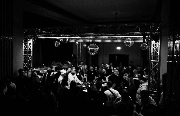 Zu sehen ist ein schwarz-weiß Foto. In der Mitte steht agieren die DJs und die Gäste tanzen um sie herum. Discokugeln sorgen für atmosphärisches Licht.