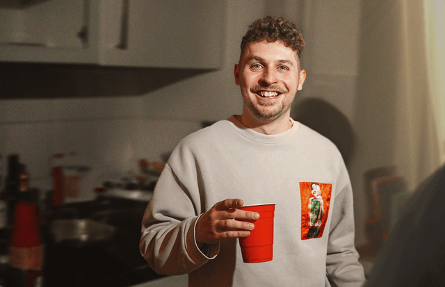 Der Comedian Fabian Lampert blickt lachend in die Kamera. In der rechten Hand hält er einen roten Plastikbecher. Der Hintergrund ist unscharf, man erahnt eine Küche.