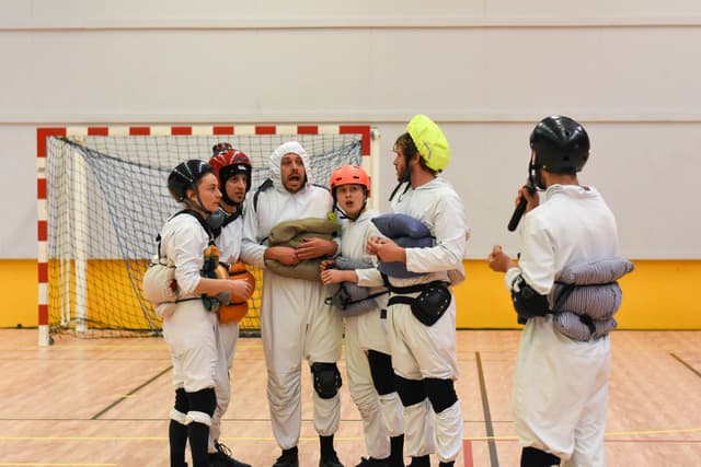 Theaterszene in einer Sporthalle: Zwei Männer in Helmen und weißen Overalls, einer davon mit mit Kissen und Polstern behängt.