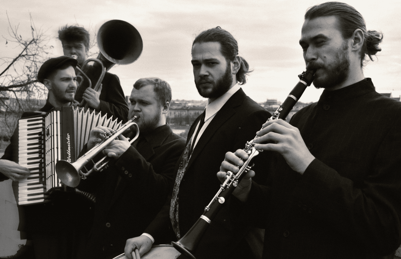 Schwarz-weiß Fotografie mit fünf Musikern in dunkler Kleidung mit ihren Instrumenten: Klarinette, Trommel, Trompete, Akkordeon und Tuba.
