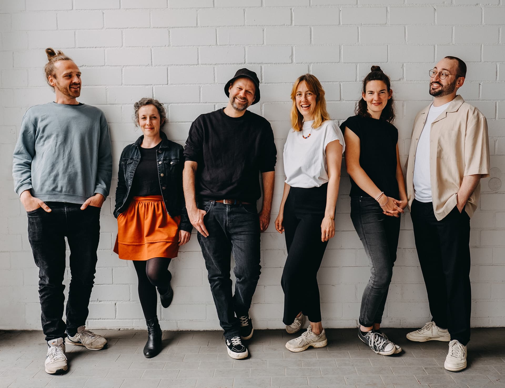 Die sechs Bandmitglieder stehen nebeneinander vor einer weißen Mauer und lachen freudig.