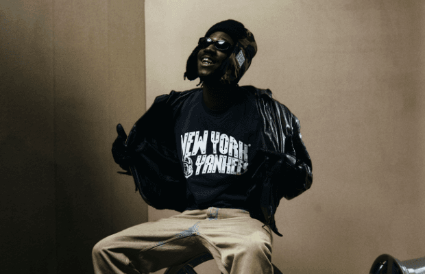 Der Hip Hop Musiker KWAM.E sitzt vor eine. beigen Hintergrund, er trägt ein dunkelblaues Shirt mit der weißen Aufschrift New York Yankees, eine schwarze Lederjacke und Lederhandschuhe. Er trägt eine Mütze mit Ohrlaschen und eine Sonnenbrille und lächelt mit Goldzähnen an der Kamera vorbei nach schräg oben.