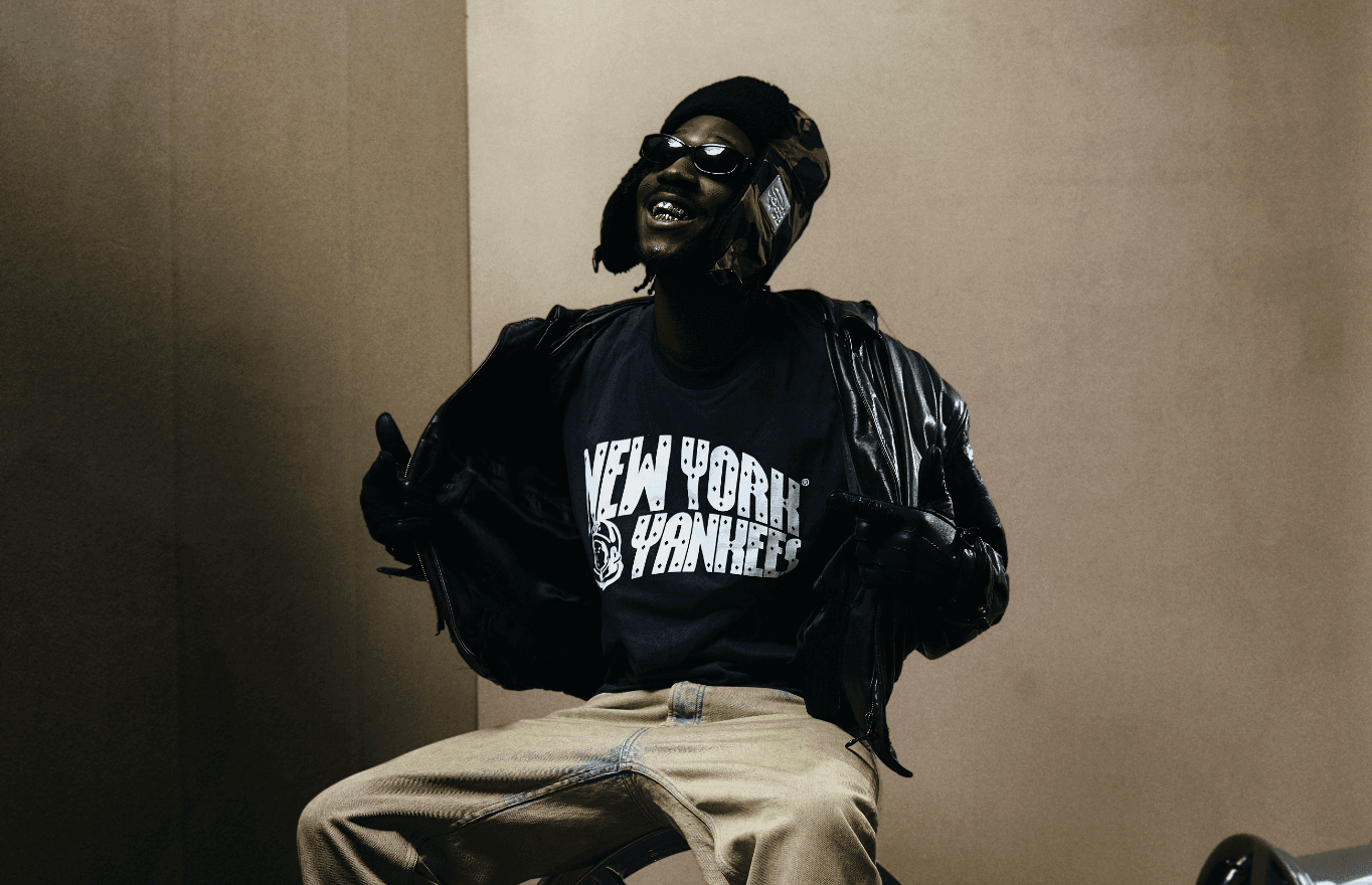 Der Hip Hop Musiker KWAM.E sitzt vor eine. beigen Hintergrund, er trägt ein dunkelblaues Shirt mit der weißen Aufschrift New York Yankees, eine schwarze Lederjacke und Lederhandschuhe. Er trägt eine Mütze mit Ohrlaschen und eine Sonnenbrille und lächelt mit Goldzähnen an der Kamera vorbei nach schräg oben.
