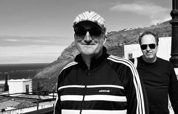 Schwarz-weiß Fotografie von zwei Männern mit Land und Meer im Hintergrund. Der eine Mann im Vordergrund trägt ein scharz-weiß gestreiftes Oberteil, eine Sonnenbrille und eine helle Kappe, der Mann rechts dahinter ein schwarzes Oberteil und Sonnenbrille.