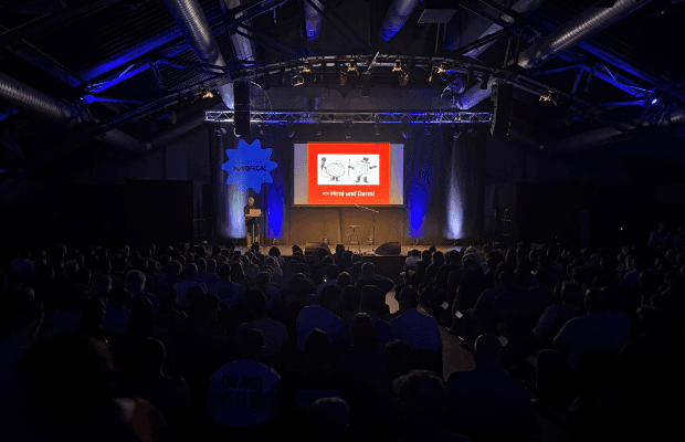 Foto von hinten in den abgedunkelten Saal der Centralstation über das Publikum hinweg auf die Bühne. Ein Redner links im Bild am Pult, im Bühnehintergrund eine rote Leinwand mit der Projektion einer Präsentationsfolie.