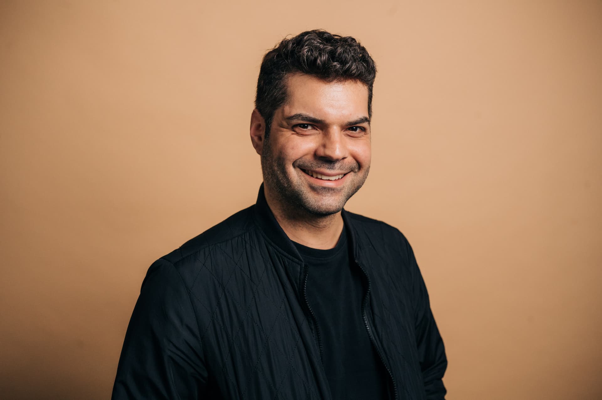 Porträtfoto des Comedian Joe Di Nardo in dunklem Shirt und dunkler Jacke vor einem beige-braunen Hintergrund, der freundlich in die Kamera lächelt.