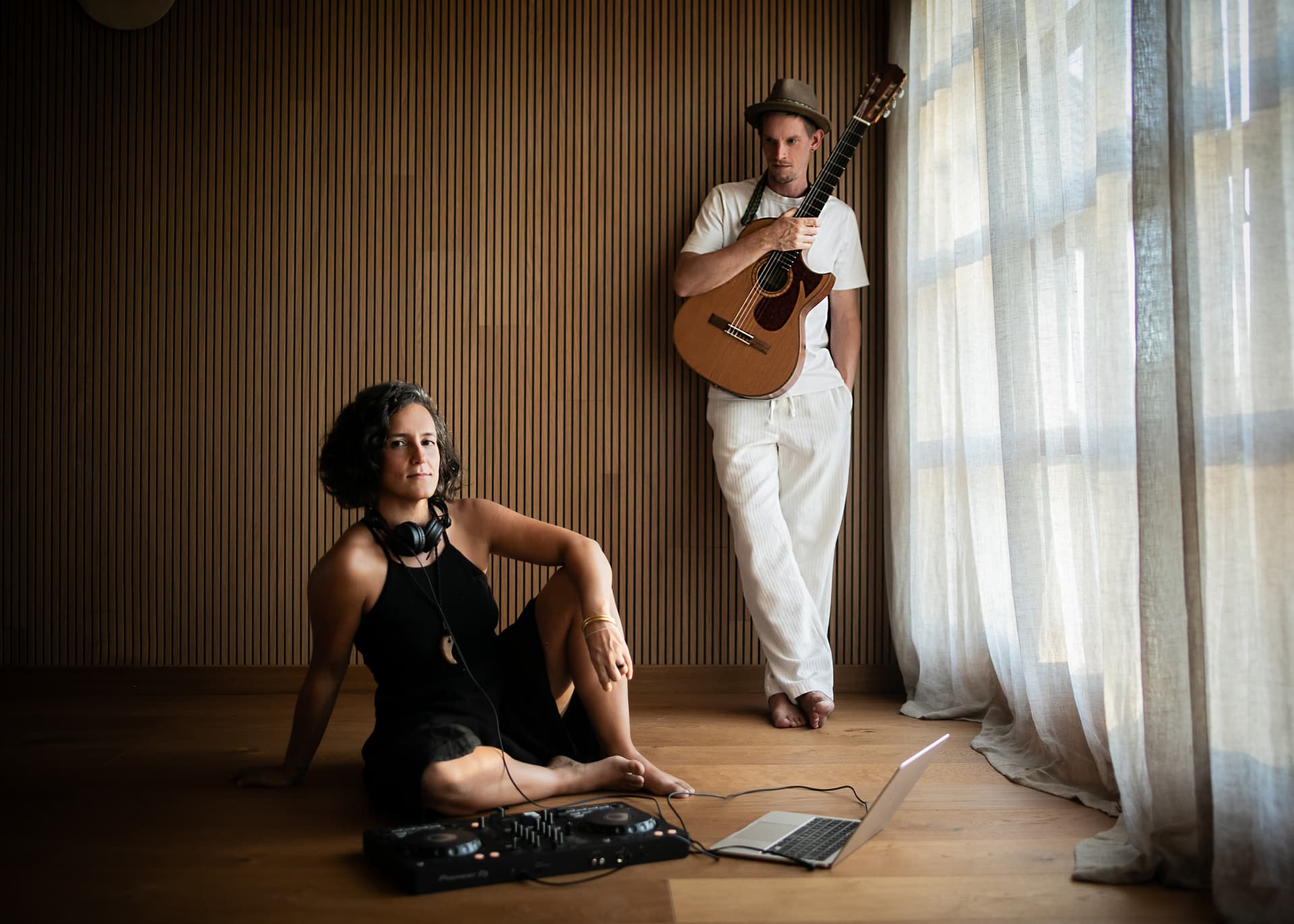 Eine Frau mit Kopfhörern und DJ-Equipment sitzt auf dem Boden, während ein Mann mit Gitarre neben ihr steht. Beide sind vor einer Holzlamellenwand in einem lichtdurchfluteten Raum zu sehen.