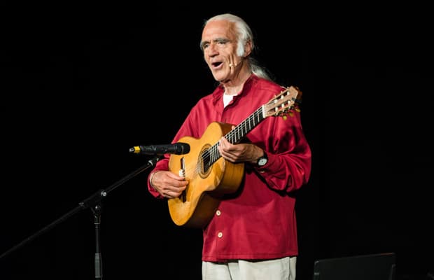 Der Musiker Fredrik Vahle steht mit einer Gitarre und mit rotem Hemd bekleidet vor einem Standmikrofon und hat den Mund wie beim Singen leicht geöffnet.