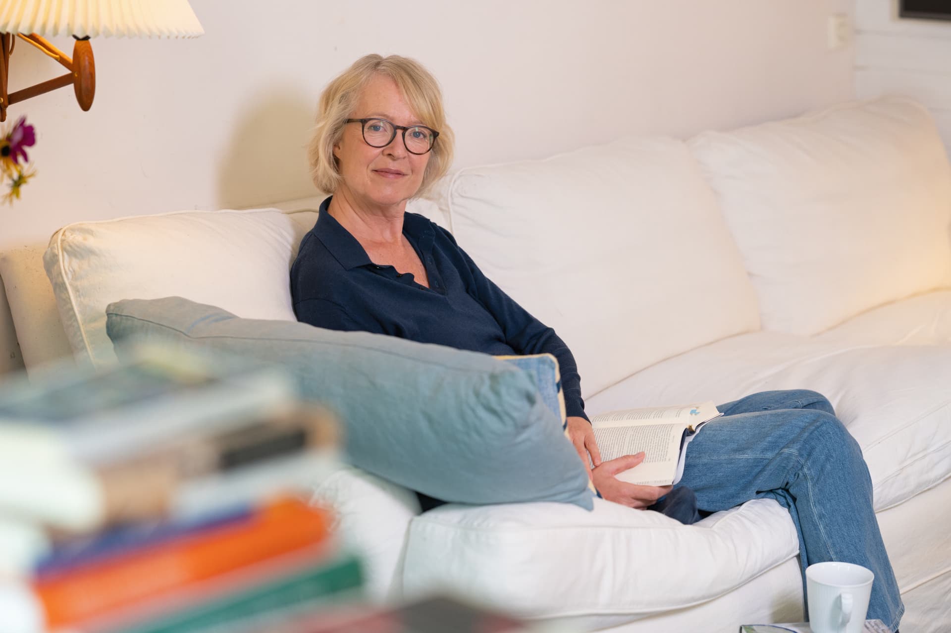 Die Autorin sitzt auf einem hellen Sofa mit einem aufgeschlagenen Buch in der Hand, sie trägt eine blaue Bluse und eine Brille und schaut seitlich lächelnd in die Kamera.