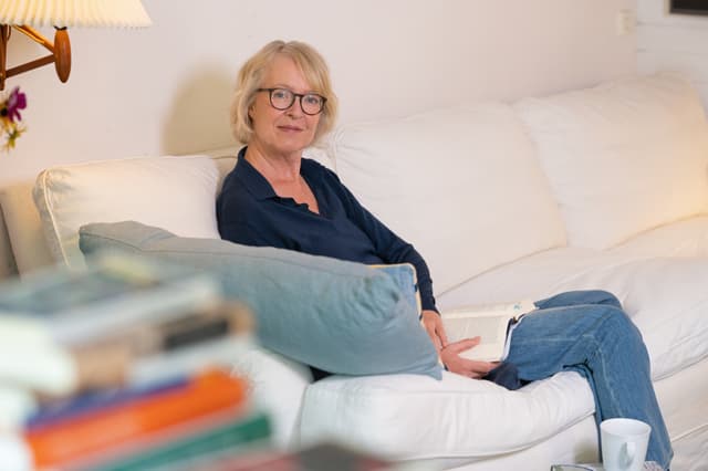 Die Autorin sitzt auf einem hellen Sofa mit einem aufgeschlagenen Buch in der Hand, sie trägt eine blaue Bluse und eine Brille und schaut seitlich lächelnd in die Kamera.